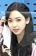 Woman with long, dark hair sitting in a recording studio. She is holding a white face mask in one hand and is smiling gently toward the camera. A studio microphone is visible in front of her, and a transparent divider separates her from the camera, reflecting parts of the room.