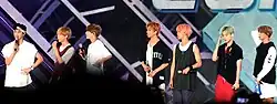 Seven men standing on a stage with each holding a microphone and dressed in casual, colorful outfits, with hairstyles varying from natural to brightly dyed colors like pink and green.