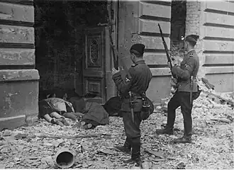 Two uniformed men holding rifles standing in front of a doorway amid rubble and debris. Inside the doorway are multiple bodies.