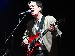 The band's frontman, performing lead vocals at the microphone, plays guitar while he sings. His face communicates strong emotion, and his posture shows great concentration on his vocal delivery.