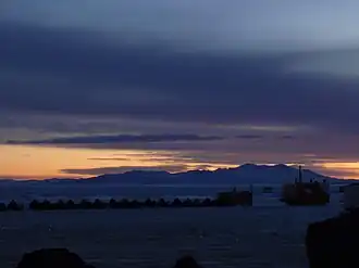 An image of Black Island. In the foreground is a Skidoo brand snowmobile. In the background is a mountain range with a sunset behind it. Above, the sky is cloudy.