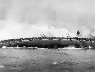 A sinking gray warship rolls over onto is side, its guns pointed skyward.