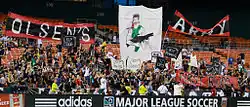 Fans wearing black cheer with several large graphics in a stadium's bleachers.