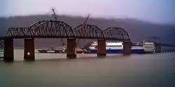 The bridge has 6 spans; the 5th has been removed from the bridge. Underneath, the ship Delta Mariner is three quarters of the way through the bridge, with the destroyed 5th bridge span draped over its front. Two boats with cranes sit at the bow of the Delta Mariner.