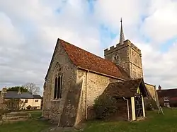 A stone church seen from the south, with a central battlemented tower, the nave with a porch and red tiled roof to the left, and a smaller chancel with a flat copper roof to the right