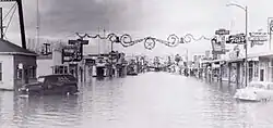 A wide street largely underwater, with old cars and buildings lining the street. The photo is black-and-white.