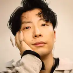 The cover art to the digital-only "Fushigi" single. Above a biege background, Hoshino – in off-white and black knitted clothing, a silver-colored ring, and turqoise-colored nails – rests his right cheek on his hand as he looks into the camera.