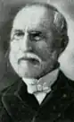 Head shot of balding gentleman with neatly trimmed white hair, mustache and beard, wearing glasses