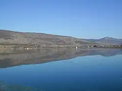 A calm, clear lake stretches across the foreground, perfectly reflecting the rolling hills and sparse vegetation on the opposite shore. The hills are covered with dry grass and patches of leafless or brownish shrubs, indicating a late autumn or early spring season. Scattered along the shoreline are small houses and cabins, some painted white or red, adding dots of color to the otherwise muted landscape. In the distance, the hills rise gently, with a few rocky outcrops and a higher peak visible on the right side of the image. The sky above is clear and bright blue, with a faint, pale moon visible high in the sky. The overall scene is tranquil and expansive, with the still water creating a mirror-like effect that doubles the landscape.