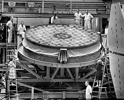 A black and white photograph of a large, flat mirror with a geometric pattern, supported on a metal frame with several people surrounding it.