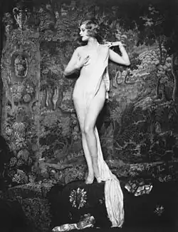 Nude portrait of a woman with brown hair looking to the side, posed in front of a tapestry