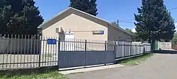 A one-story beige building with a grey metal fence and gate. It is surrounded by green trees under a clear blue sky.