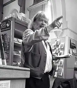 A photograph of a rotund bearded jolly man pointing to the audience with one hand while reading from a book of his poetry with his other.