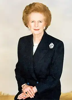 Half-length portrait of Thatcher. She is wearing a black suit and pearls, with a brooch on her left lapel. Her hands are clasped in front of her, adorned with rings. The background is pale and softly lit.