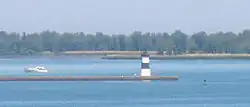 A pier with a square, black and white-striped lighthouse at the end. A small motorboat is paralleling the pier, heading away from the lighthouse.