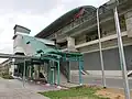 A photograph of Teck Lee station's exterior. It showcase one of the station's exits, an overhead pedestrian bridge leading to the side of a road. The station, which is on the centre of a road, has a boat-shaped roof. There is a rail viaduct that leads to the station