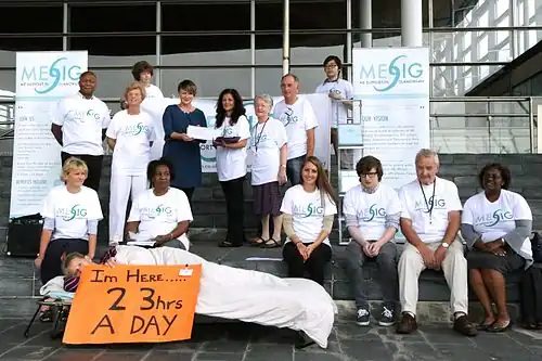 a group of people offering a petition. The group includes a person on a camp bed holding up a placard that says she is in bed 23 hours a day.