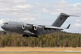 Colour photo of a grey military aircraft flying just above the ground with its wheels extended