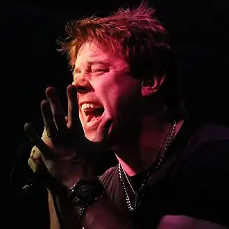 close-up of Shimon Moore lit up from the front in a dark background, singing/screaming into a microphone
