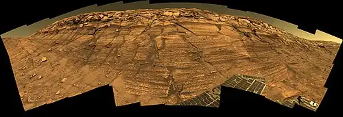 Figure 6. Burns Cliff in Endurance Crater. This image is one of the best known by Opportunitys Pancam. The original file is a huge (12,316 x 4,218 pixels) "mosaic" of many images. It has a "fish eye" view distortion. Click on the image for higher resolution. Burns Cliff is at least 7 m tall. Meridiani sediment cliffs as big as Burns Cliff are not common. They only occur in large Meridiani craters younger than the sediments. The sediments in Burns Cliff are more weathered than those in Figure 5 since Endurance Crater is much older than Concepcion Crater. Opportunity collected data at Burns Cliff over several months in 2004, and many important papers used this data. Images taken on Sols 287-294 (2004-11-13 to 2004-11-20).