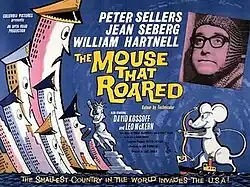 A cartoon cityscape targeted by a mouse with a bow and arrow. Inset is a photograph of a glum looking man wearing chain mail armor and thick black glasses.