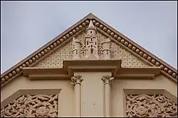Close-up of a castle sculpture situated below the apex of a roof. The sculpture and surrounding wall are all painted the same pastel-yellow.