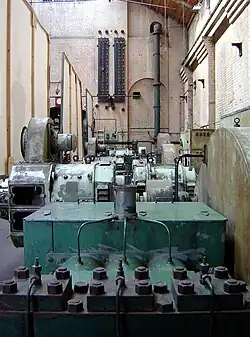Large, dark machinery and pipes dominate a tall, brick-walled space with windows.