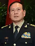 a smiling man with his mouth open and looking rightwards, wearing a light green military uniform with insignia and badges